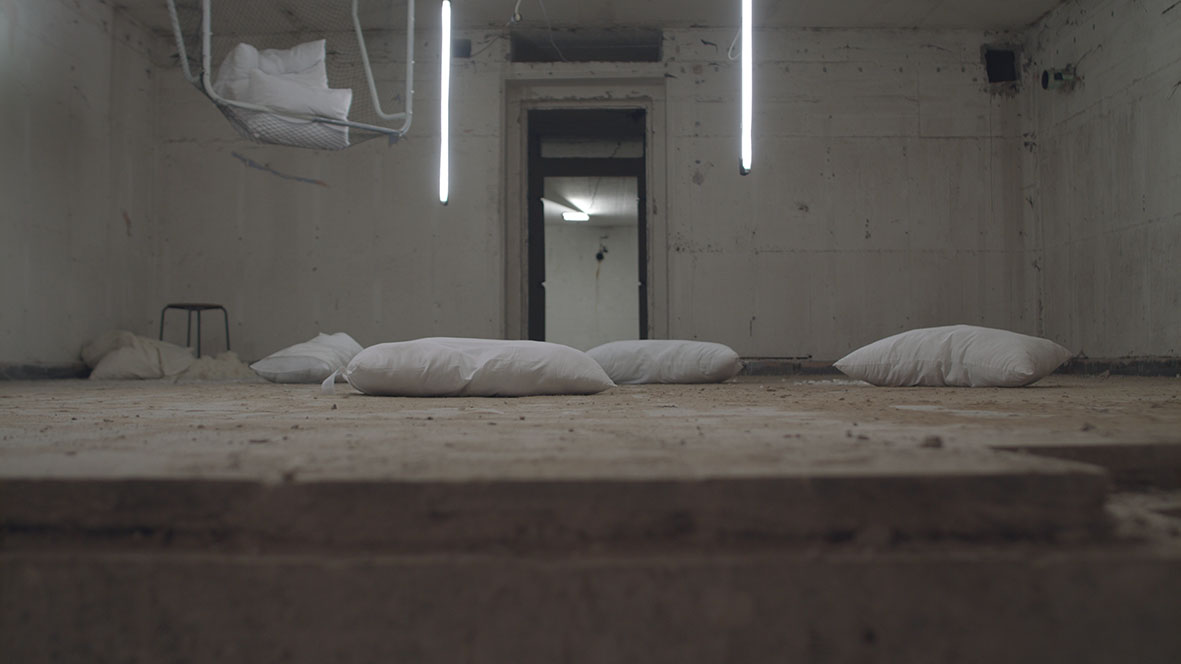 A coloured video still of a room with white pillows scattered all over the floor. Tow fluorescent strip light hanging vertically off the ceiling. A stool in the left-hand corner of frames with feathers on th ground around it. A small icehockey goal post is haning upset from the ceiling with a couple of white pillows caught in it. In the middle of the room is a door with a view inside another room with fluorescent lighting